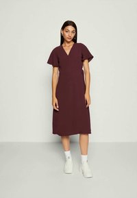 Mulher de pé, vestida com um vestido envolvente curto de manga curta, na altura do joelho, na cor bordô, usando meias brancas e tênis brancos robustos, contra um fundo simples.