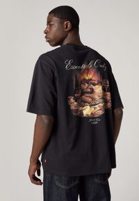 T-shirt de algodão preta com uma imagem texturizada de comida nas costas, com o texto "Essentials Only" acima. Corte casual, mangas curtas.
