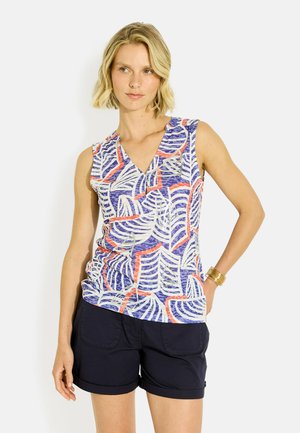 Mujer que lleva una camiseta sin mangas con patrones de hojas en azul, blanco y naranja y pantalones cortos negros, posando con una mano en la cadera.