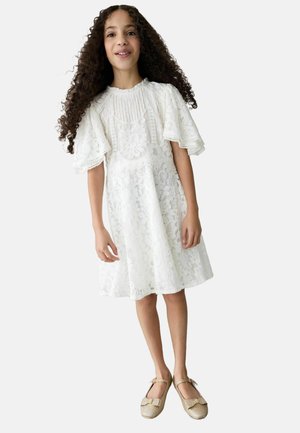 Weißes Spitzenkleid mit kurzen, Rüschenärmeln, das mit Blumenmustern verziert ist. Kombiniert mit glitzernden, goldenen Ballettschuhen und einer schlichten Silhouette.