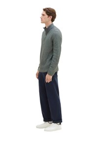 Grauer Reißverschluss-Pullover mit strukturierter Oberfläche, kombiniert mit marineblauen, weit geschnittenen Hosen und weißen Sneakers, seitliche Ansicht vor einem weißen Hintergrund.