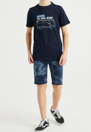 Boy wearing navy graphic T-shirt with van and "Living on the Road" text, blue tie-dye shorts, and black-and-white sneakers walking.