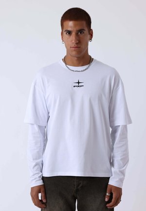 Jeune homme aux cheveux roux courts portant des chemises blanches superposées à manches longues et courtes avec le logo "PXP", chaîne en argent, boucles d'oreilles et bagues, debout devant un fond blanc.