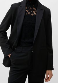 Blazer nero con rever a punta, realizzato in tessuto morbido. Indossato sopra un top in pizzo nero, con collo alto e motivi intricati.