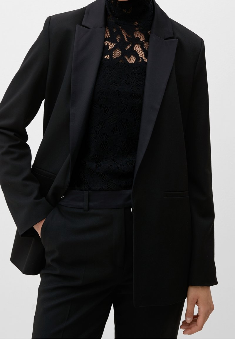Blazer nero con rever a punta, realizzato in tessuto morbido. Indossato sopra un top in pizzo nero, con collo alto e motivi intricati.