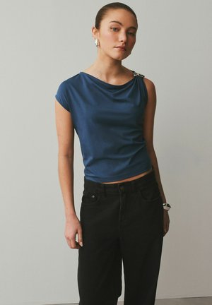 Tânără cu părul prins la spate, purtând un top albastru fără mâneci și pantaloni negri cu talie înaltă, stând în fața unui perete simplu, deschis la culoare.