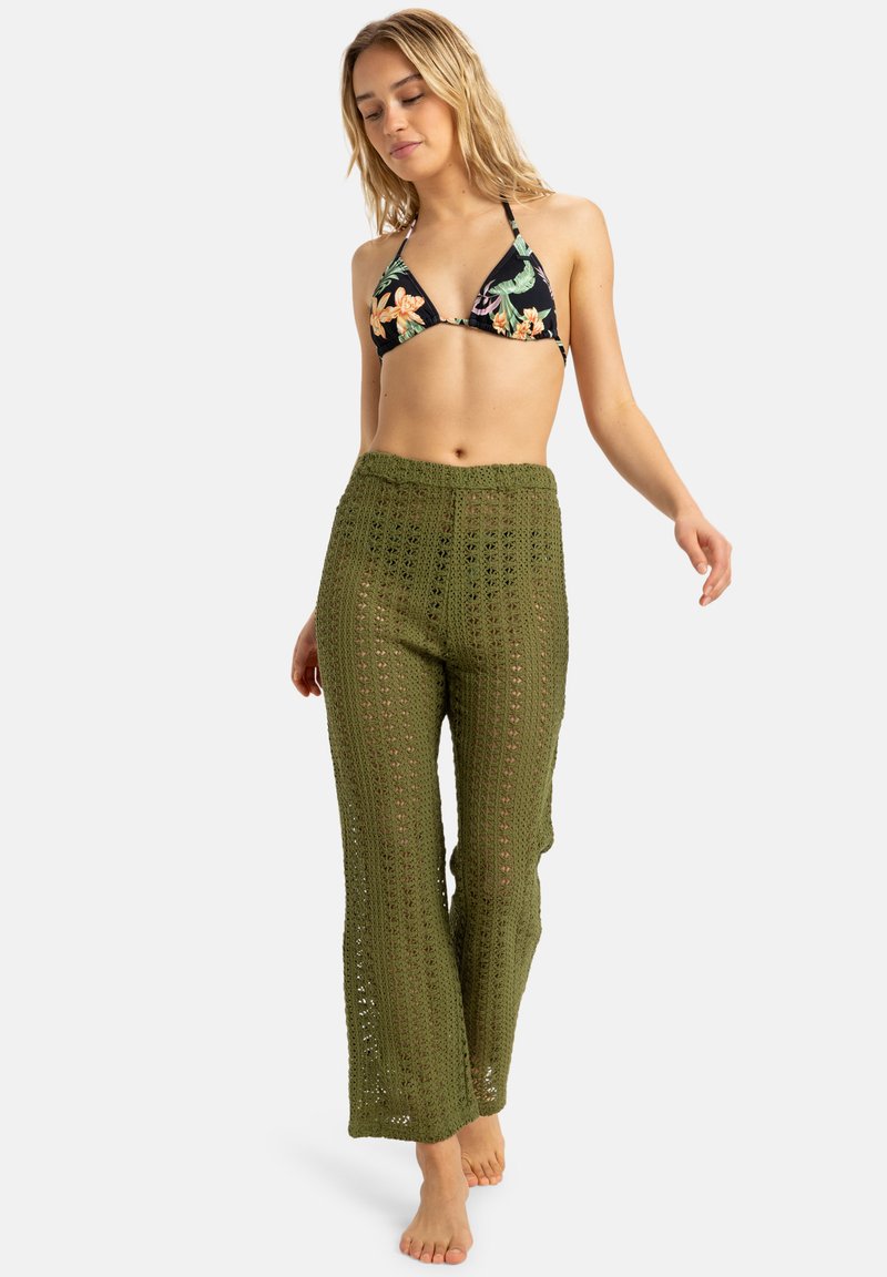 Crochet, olive green pants with an open weave pattern, paired with a black floral bikini top. Model walking barefoot, casual fit.