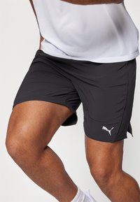 Sorte atletiske shorts lavet af letvægtigt stof med sideskår, der har et hvidt Puma-logo på den nederste venstre ben.