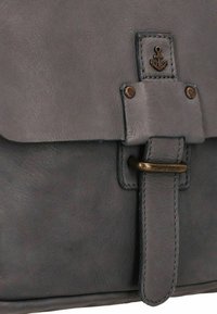Borsa in pelle grigia con una texture liscia, dotata di una patta frontale, accessori in bronzo e un emblema ad ancora cucito sulla chiusura.