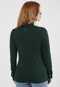 Chemise à manches longues vert foncé avec un col haut, présentant des poignets boutonnés et un détail de logo au dos. Tissu lisse et extensible.