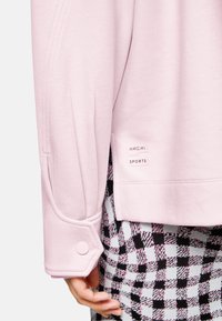 Light pink sweatshirt with a buttoned cuff, featuring seams and a logo tag in white. Textured, patterned fabric visible underneath.