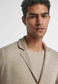 Beige Strickblazer mit weicher Textur und Schalkragen, getragen über einem passenden hellbeigen T-Shirt mit minimalistischem Design.