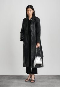 Lungo cappotto di pelle nera con chiusura a bottoni, indossato sopra un top e pantaloni neri. Borsa argentata con dettagli in frange. La modella è in piedi su un pavimento grigio.