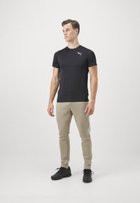 Muž stojící v černém tričku Puma, béžových teplácích s bílými postranními pruhy a černých teniskách na bílém pozadí.