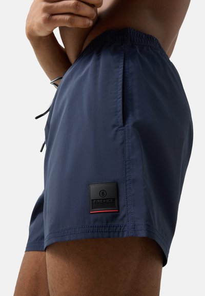 Mand iført marineblå badeshorts med elastisk talje og sort Fire+Ice-logo på venstre lår mod en ensfarvet baggrund.