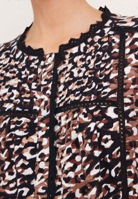 Haut à motifs léopard avec texture plissée, bordure en dentelle noire au niveau du col et de la poche, avec des boutons blancs. La palette de couleurs comprend des bruns et des noirs.