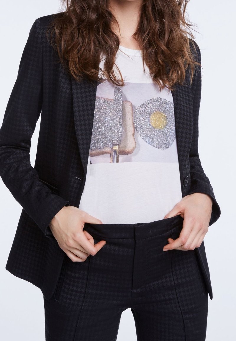 Blazer nero con texture e motivo a pied de poule, indossato sopra una t-shirt bianca con grafica scintillante, abbinato a pantaloni neri.