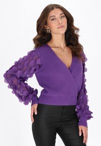 Suéter cruzado morado con tela translúcida texturizada en las mangas, con un patrón de panal y cuerpo de punto acanalado; combinado con pantalones negros.