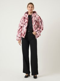 Mulher vestindo uma jaqueta acolchoada com padrão rosa e vermelho sobre uma blusa preta e calças largas pretas, de pé contra um fundo claro e simples.