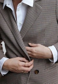 Blazer à motif pied-de-poule marron et blanc avec un revers cranté et une fermeture à un bouton, porté avec une chemise blanche à col en dessous.