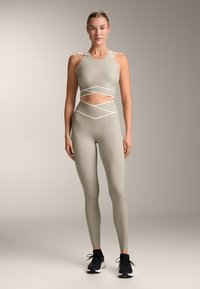 Bež športni nedrček in legging set s perforiranim dizajnom, tankimi kontrastnimi naramnicami in gladkim, elastičnim materialom. Nosijo se črne športne čevlje.