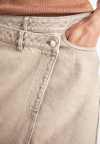 Un pantalon en denim beige présente une coupe classique avec une fermeture à bouton et une poche latérale. Le tissu a une finition texturée avec des coutures contrastantes.