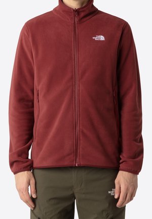 Mand iført en mørkerød fleecejakke med lynlås og olivengrønne bukser, begge med The North Face-logo.
