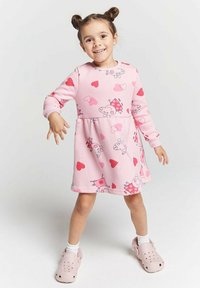 Lächelndes junges Mädchen mit zwei Duttfrisuren in einem rosa Kleid mit Cartoon-Schweinen und Herzen, trägt weiße Socken und rosa Slipper, stehend vor weißem Hintergrund.