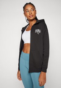 Sudadera con cremallera negra con bolsillos frontales, logotipo bordado de "PUMA sports club", tela suave, combinada con una camiseta blanca y leggings azules.