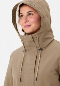 Donna che indossa una giacca con cappuccio beige foderata in pile, guarda leggermente verso l'alto e sorride dolcemente.