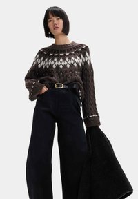 Maglione marrone lavorato a maglia con motivi bianchi e scuri, abbinato a pantaloni neri a vita alta. Il modello tiene in mano una giacca nera con texture.