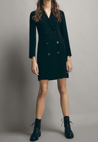 Schwarzes, doppelt-reihiges Kleid mit langen Ärmeln, ausgestattet mit zwei Vordertaschen und großen Knöpfen. Kombiniert mit schwarzen schnürbaren Stiefeln im Combat-Stil.