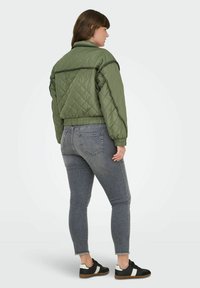Padded olivno zeleni bomber jakna s črnim obrobljanjem, z elastičnimi manšetami in pasom, nosi se čez sivo jeans hlače in črne superge.