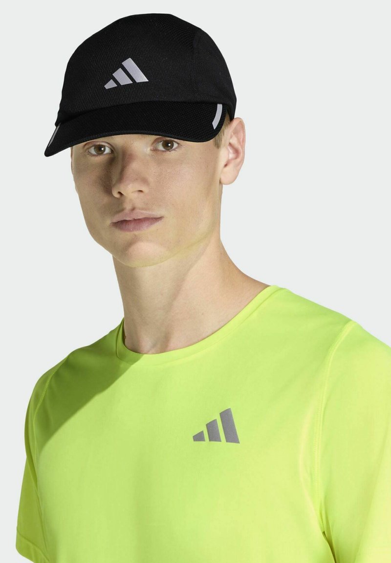 Gorra deportiva negra con diseño de malla y acentos grises. Se lleva con una camiseta de color verde neón brillante que presenta un discreto detalle de logo.