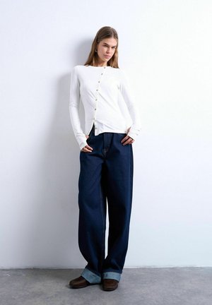 Kvinde iført en hvid asymmetrisk bluse med knapper, brede mørkeblå bukser med opbuk ved manchetterne og brune sko, stående op ad en hvid væg.