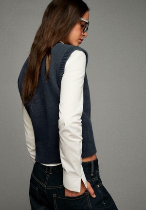 Donkerblauw gebreid vest met geribbelde randen over een witte lange mouw shirt, gecombineerd met donkere spijkerbroek; gladde texturen en gelaagde look.