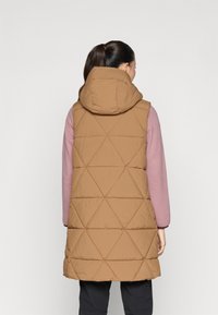 Gilet trapuntato marrone con cappuccio, caratterizzato da motivi a diamante. Indossato sopra una maglietta rosa a maniche lunghe, dal taglio semplice e dalla texture liscia.