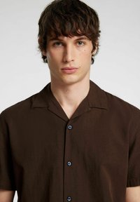 Camicetta marrone a maniche corte realizzata in un tessuto testurizzato, con colletto classico, tre bottoni blu e una vestibilità rilassata.