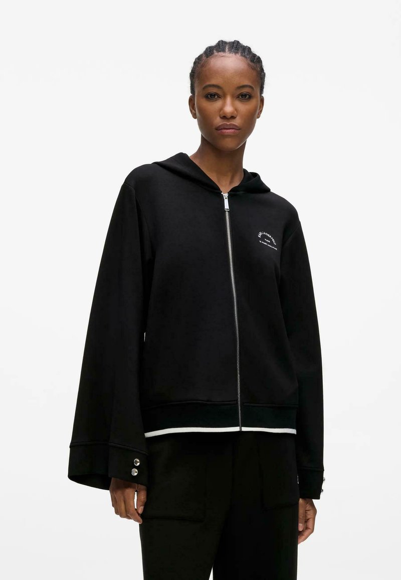 Femme portant un sweat à capuche noir zippé avec des manches larges et des détails à boutons, debout devant un fond blanc uni.