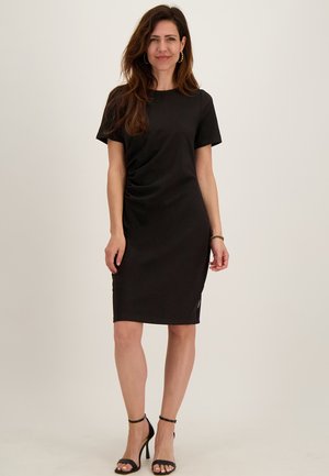 Vrouw draagt een zwarte jurk met korte mouwen tot op de knie, met zijsoufflés en zwarte hoge hakken met enkelbandje, staand tegen een witte achtergrond.