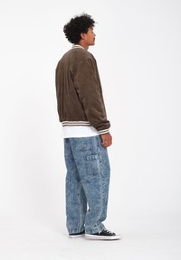 Bruine corduroy bomberjack met witte geribbelde boorden, gecombineerd met lichtgewassen baggy jeans met voorzakken en zwarte sneakers.