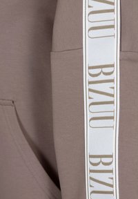 Brązowa bluza z kapturem o miękkiej teksturze, z białym paskiem po bokach, na którym znajduje się złote napisy "BIZUU" oraz kieszenią z przodu.