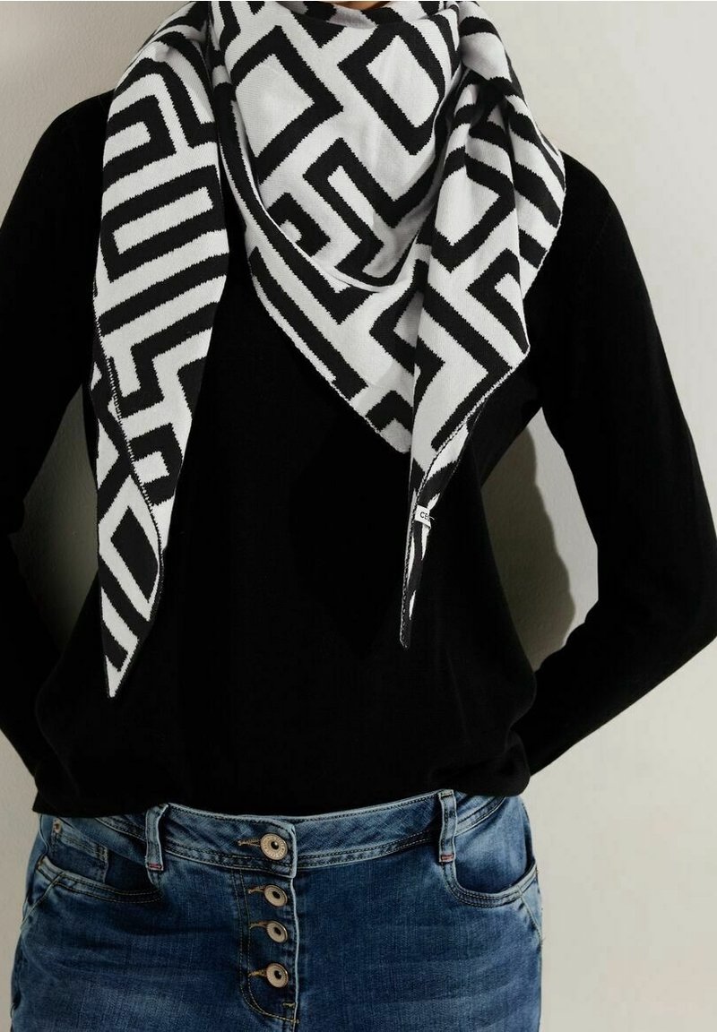 Écharpe à motif géométrique noir et blanc portée sur un haut noir et un jean bleu boutonné sur une personne avec les mains dans le dos.