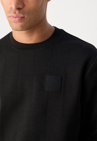 Hanorac negru cu dungi verticale și un patch texturat cu logo pe piept. Guler rotund ribflat și textură de țesătură netedă.