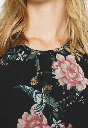 Blonde vrouw die een zwarte bloes met roze en witte bloemen en groene bladeren draagt, met focus op de halslijn en borstgebied.