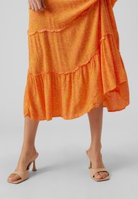 Falda de capa naranja con un patrón sutil, con detalles de dobladillo con volantes. Combinada con sandalias de tacón cuadrado abiertas en la parte delantera de color beige.