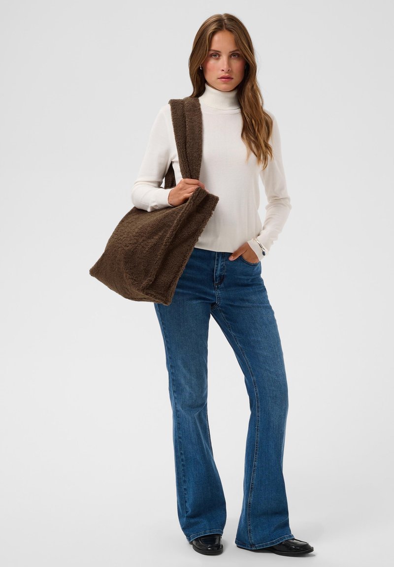 Femme portant un col roulé blanc, un jean bleu, des chaussures noires, portant un grand sac cabas marron texturé sur son épaule sur un fond uni.