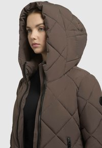 Quilted brun pufferjakke med stor hætte, frontlynlås og sidelommer. Har en struktureret finish og et cirkulært logo på ærmet.