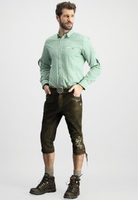 Grünes karierter Button-up-Hemd, braune Lederhosen mit Stickerei, graue Socken und dunkle Stiefel, posierend vor einem weißen Hintergrund.