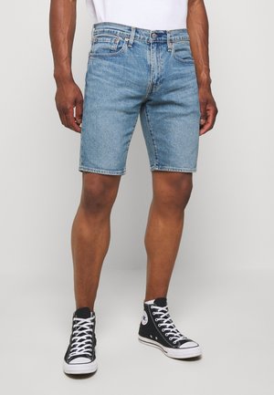 Homme portant un short en denim bleu clair, un t-shirt blanc et des baskets montantes noires avec des lacets blancs, debout devant un fond uni.